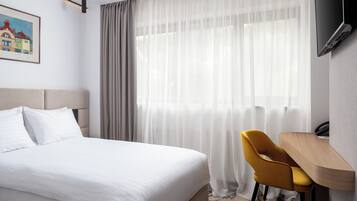 Habitación de hotel con cama, escritorio, silla, televisor y ventana con cortinas translúcidas.