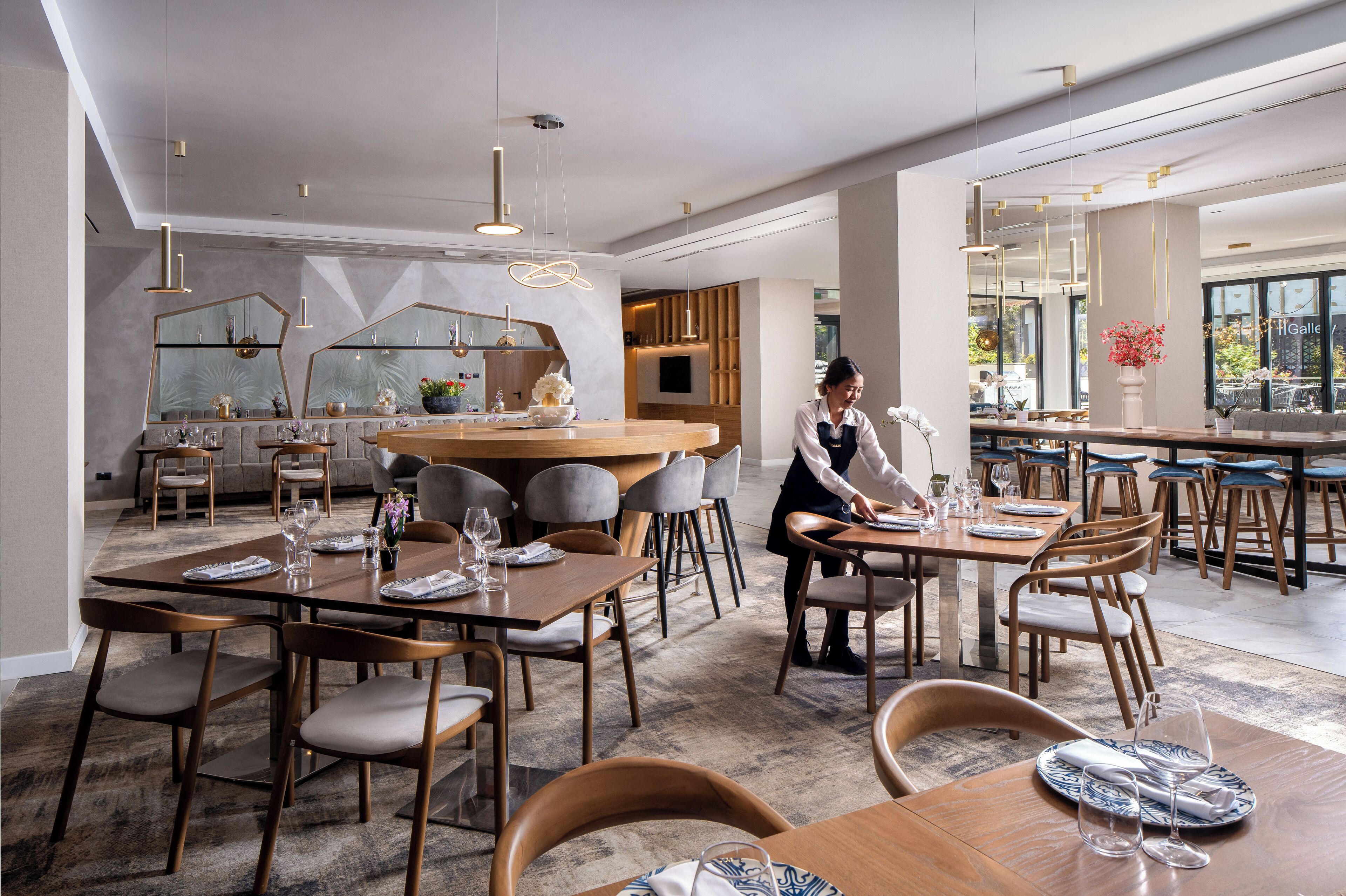 A modern restaurant with wooden tables and chairs, a central bar area, and large windows offering views of the outside.