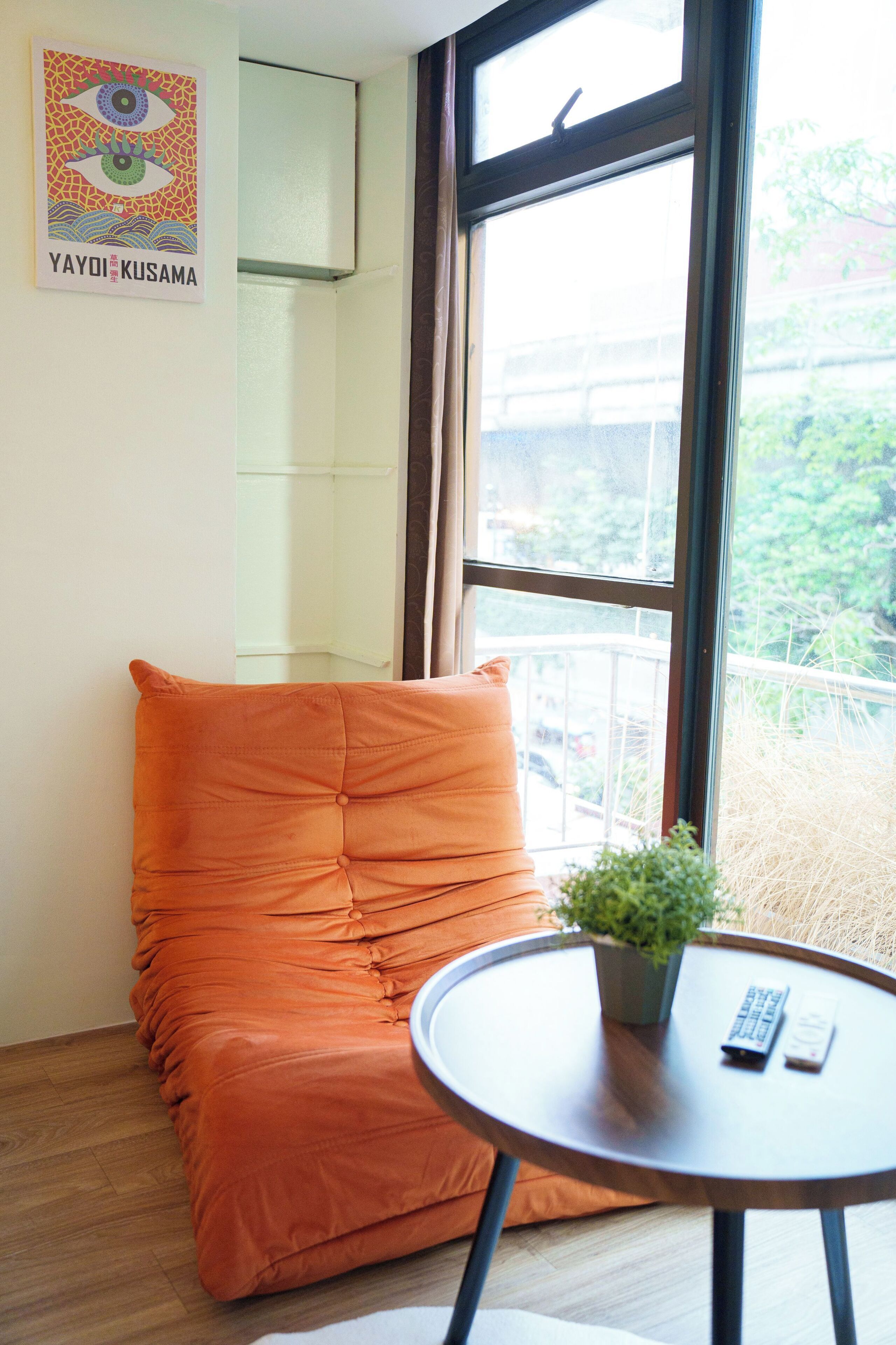 A room with a large window, a cushioned seating area, a small table with a plant, and a poster on the wall.