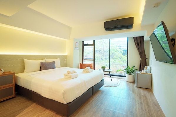 A modern hotel room with a large bed, a flat-screen TV, a small table with a plant, and a window with a view of greenery.