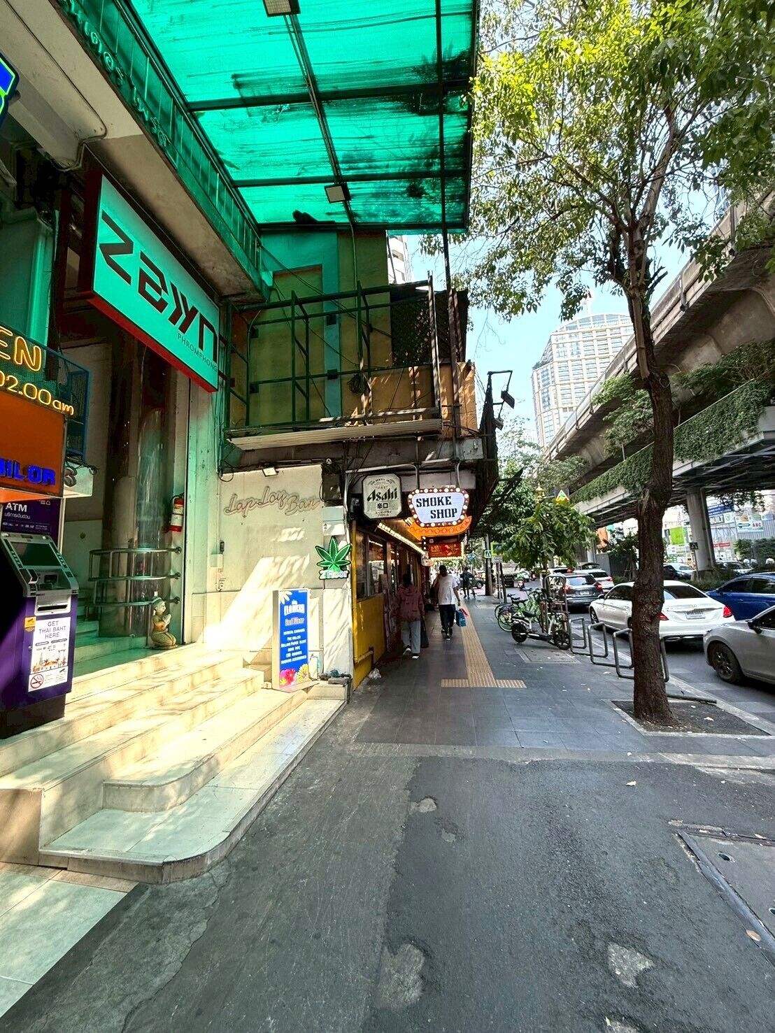A street-level view of a commercial area with a green awning, a sign for a shop named 'Asahi,' and a visible ATM.
