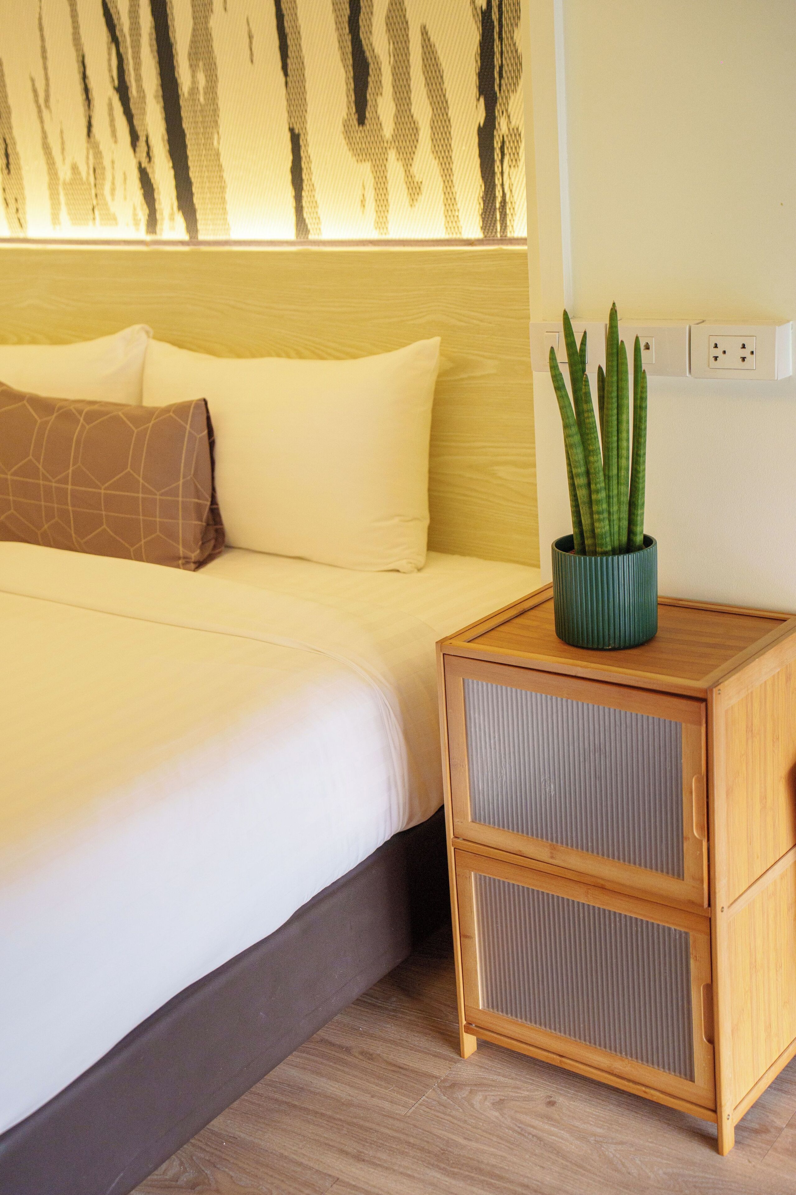 A neatly made bed with white bedding, a brown pillow, and a green plant in a vase on a wooden nightstand.