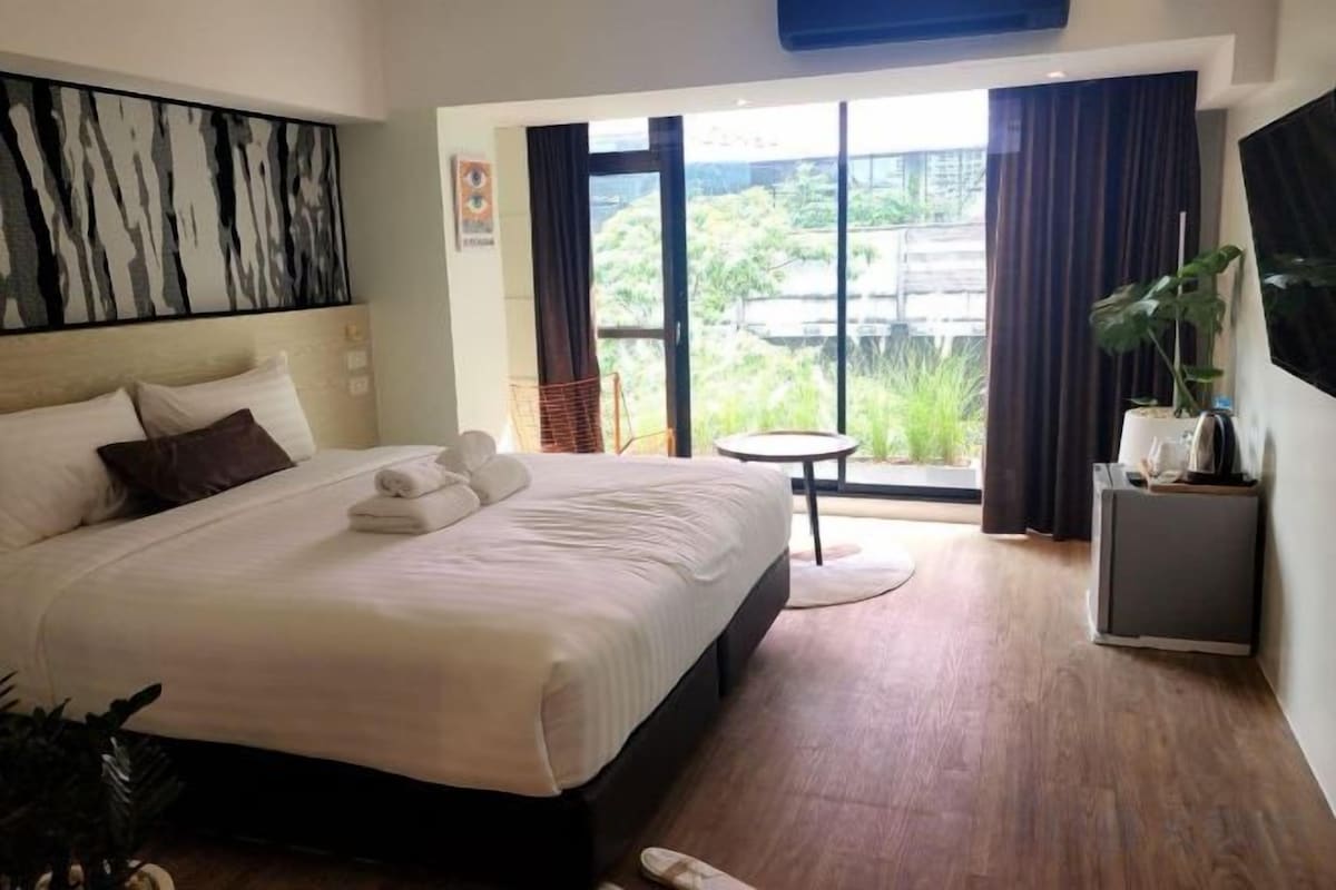 A hotel room with a large bed, a small table, a chair, and a view of greenery through the window.