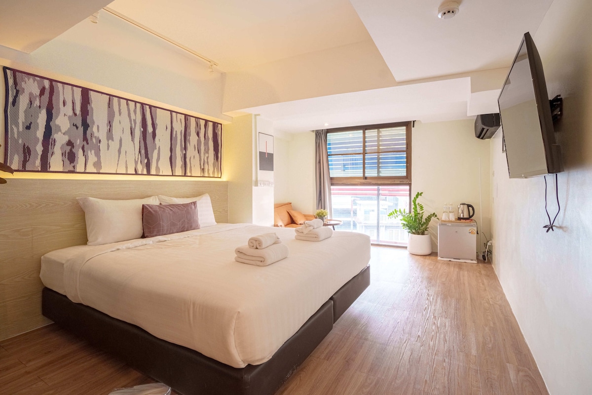 A modern hotel room with a large bed, a flat-screen TV, a small kitchenette, and a large window with blinds.
