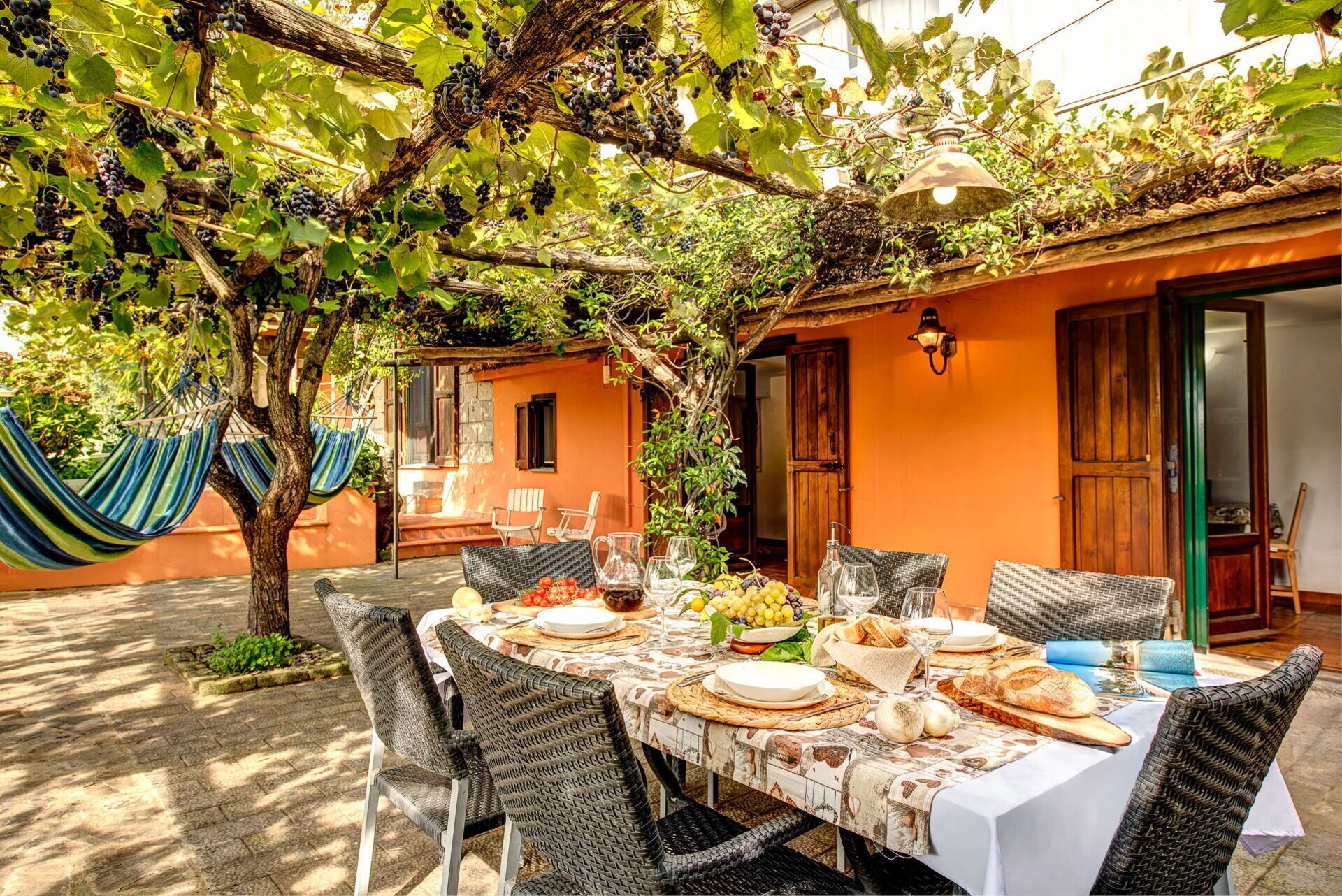 An outdoor dining area with a table set for a meal, surrounded by wicker chairs, and a hammock hanging from a tree.