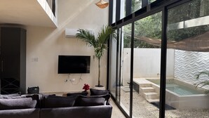 A modern living room with a flat-screen TV, a large window, and a view of a swimming pool. - VILLA ESCONDIDA - RIVIERA MAYA - LUXURY HOME WITH HIGH QUALITY DESIGN (Cancún)