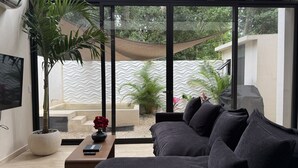 A modern living room with a sofa, a coffee table, and a TV. There are plants and a hammock outside the window. - VILLA ESCONDIDA - RIVIERA MAYA - LUXURY HOME WITH HIGH QUALITY DESIGN (Cancún)