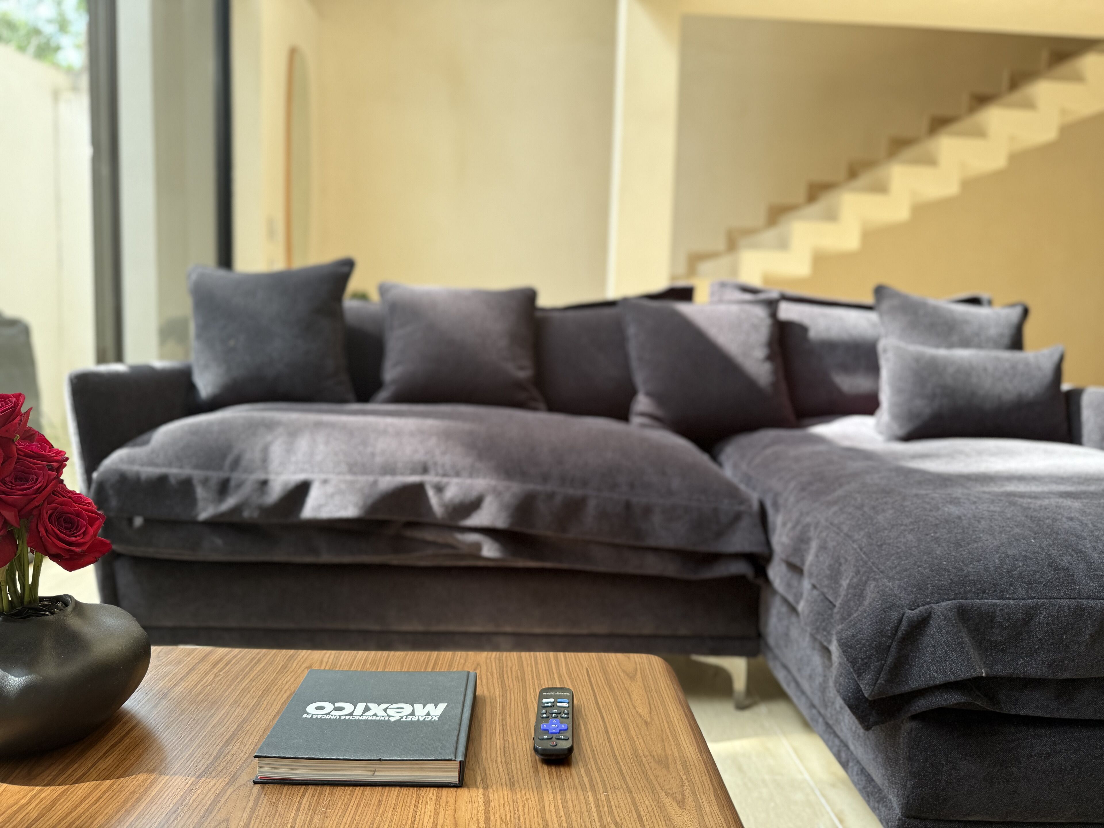 Ein modernes Wohnzimmer mit einem grauen Ecksofa, einem Couchtisch aus Holz mit Buch und Fernbedienung sowie einer Vase mit roten Rosen.