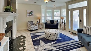 A living room with a fireplace, a ceiling fan, a sofa, a patterned ottoman, and a large window with blinds. - Corner Castle by Southern Vacation Rentals (Destin)