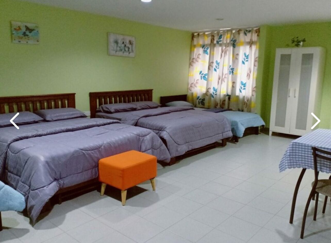 Une pièce comprenant trois lits, un petit pouf orange, une table recouverte d’une nappe à carreaux et une armoire.
