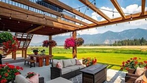 An outdoor patio with wicker furniture, a stone floor, and a view of a green field and mountains. - Luxe Barndominium - Mtn Views! "Hemlock" (Columbia Falls)