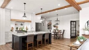 A modern kitchen with a central island, wooden beams, pendant lighting, and a dining area with a table and chairs. - Luxe Barndominium - Mtn Views! "Hemlock" (Columbia Falls)