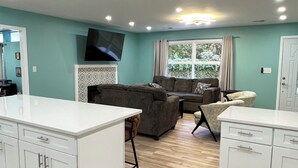 A modern kitchen with white cabinets and a central island, a grey sofa, and a television mounted on the wall. - 5 BR King Beds Game Room Yard Family Home Explore DC Quantico Stafford  (Stafford)