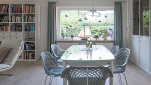 Un coin repas avec une table en verre et des chaises en fil de métal, une bibliothèque et une fenêtre donnant sur un paysage verdoyant et un voilier.