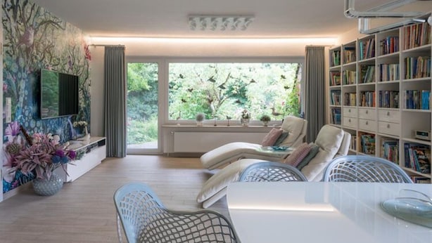 Un salon moderne comprenant un canapé, des fauteuils, une table à manger, une bibliothèque et une grande fenêtre donnant sur un paysage verdoyant.