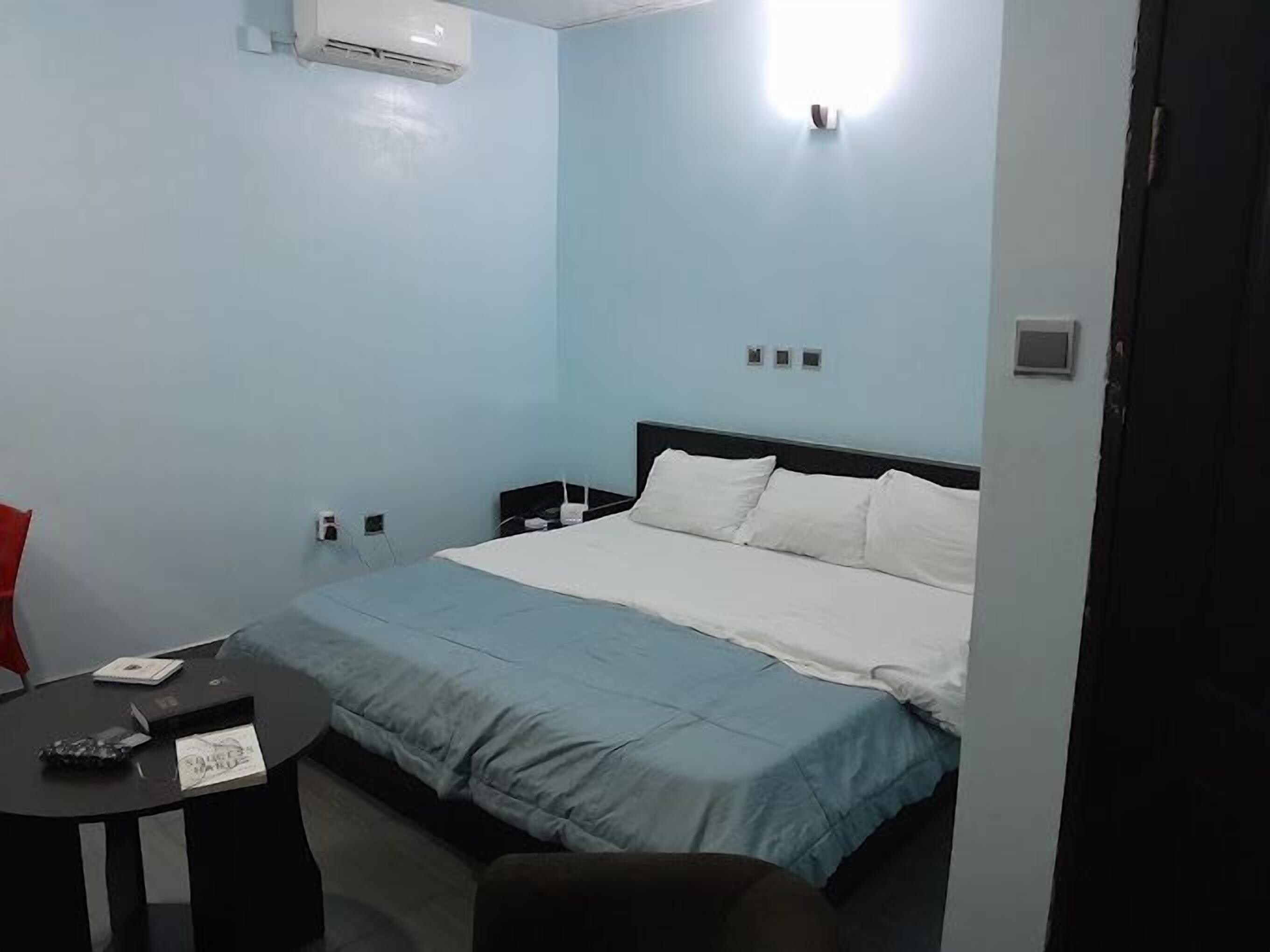 A hotel room with a single bed, a small table, and a chair. There is an air conditioning unit on the wall and a door to the right.