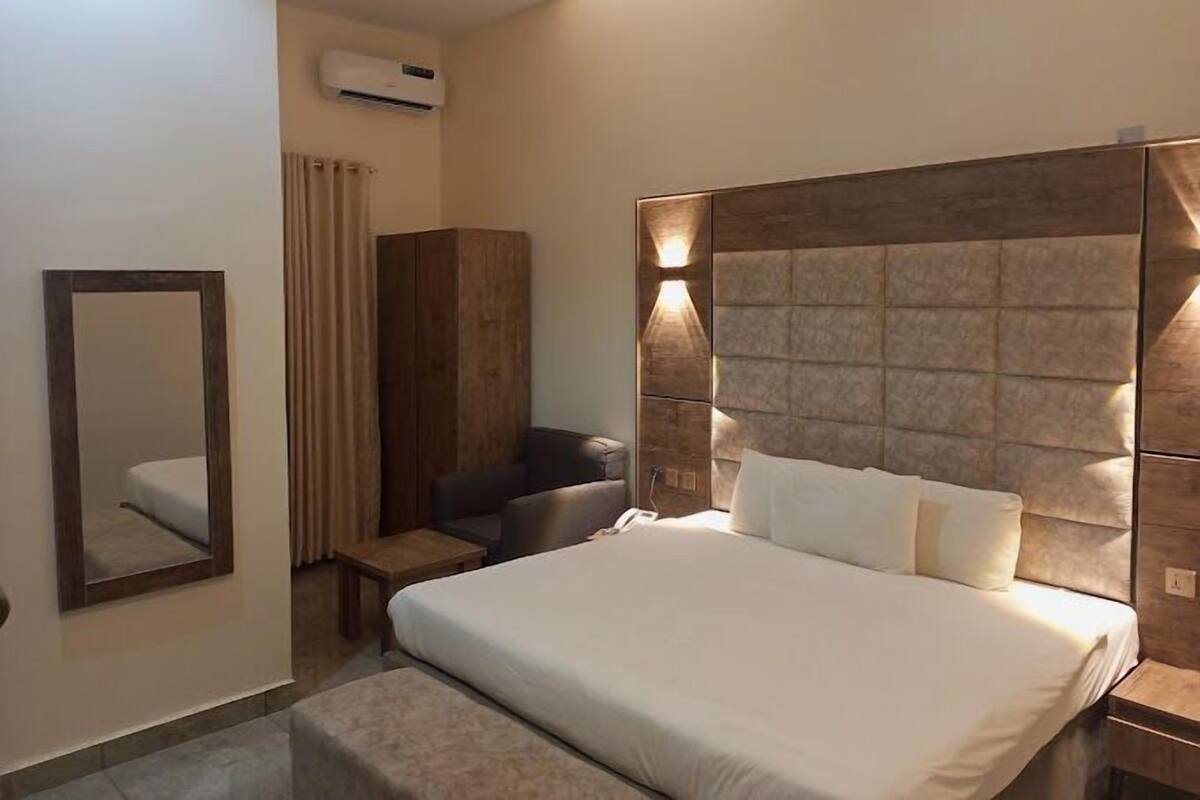 A hotel room with a large bed, a wooden wardrobe, a seating area with a chair, and a mirror.