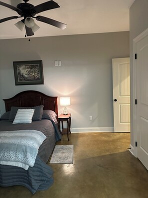 Une chambre à coucher avec un lit, une table de chevet avec une lampe, un ventilateur de plafond, un tableau encadré au mur et une porte.
