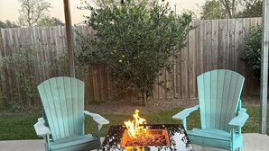 A covered patio with two turquoise Adirondack chairs, a glass-top fire pit with flames, and string lights. - Cajun Country Escape  (Gonzales)