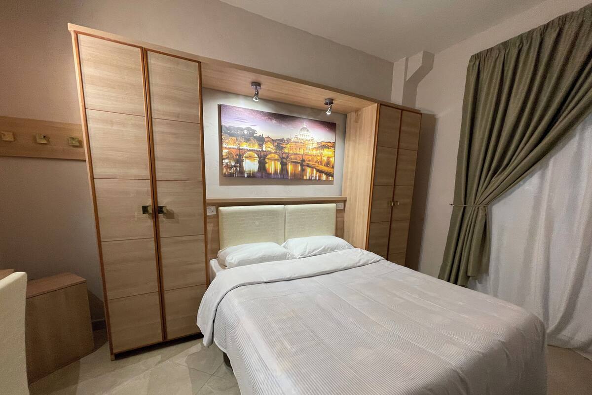 A hotel room with a bed, wooden wardrobe, and a framed picture on the wall.