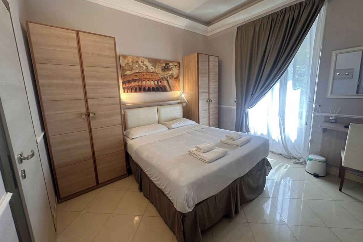 A hotel room with a large bed, a wardrobe, a painting of the Colosseum, and a window with curtains.