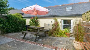 Eine Terrasse mit Picknicktisch und einem rosa Sonnenschirm, ein Haus mit Ziegeldach und Kübelpflanzen.