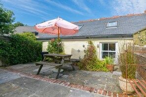 A patio with a picnic table and a pink umbrella, a house with a tiled roof, and potted plants.