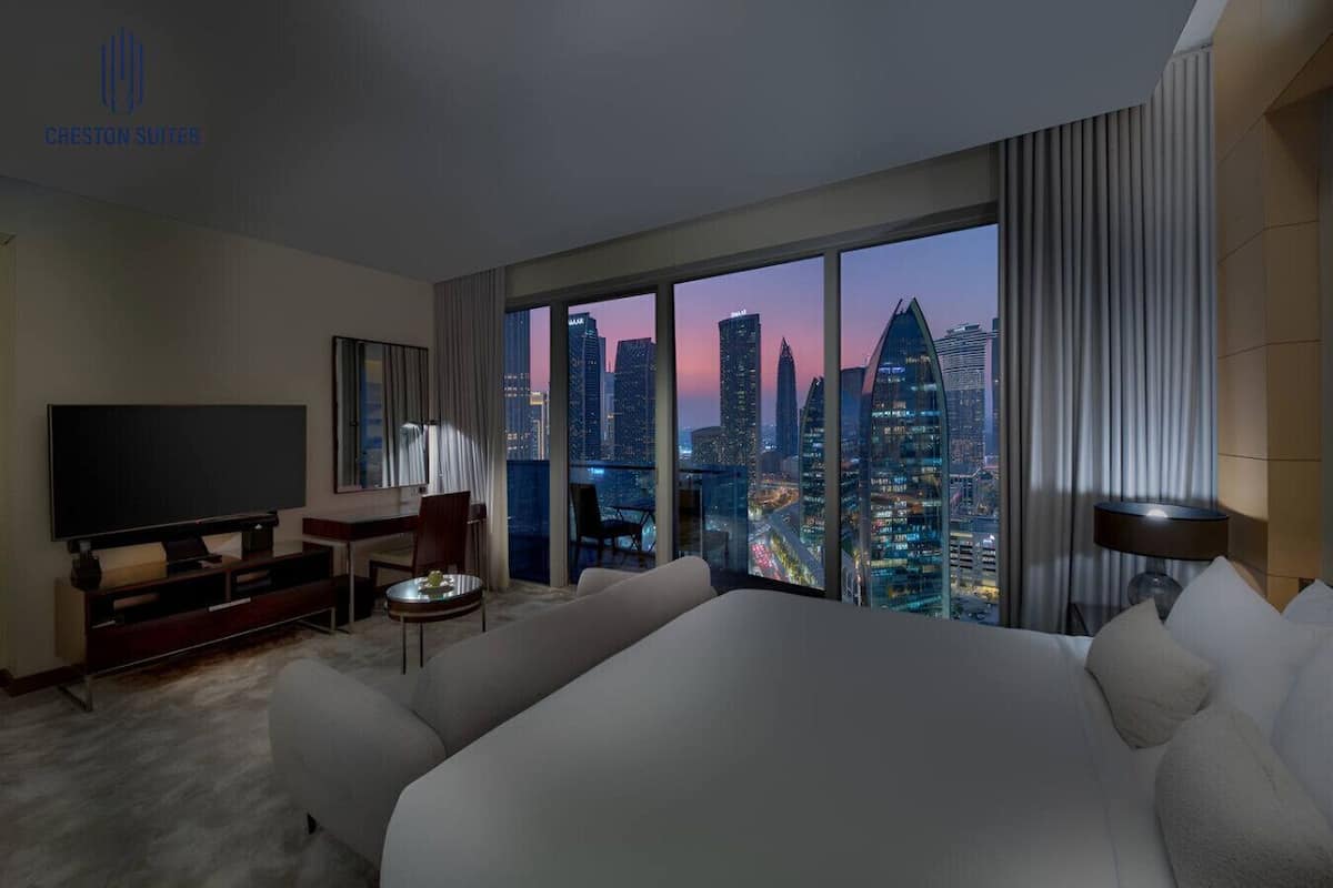 A hotel room with a large bed, a TV, a desk, and a view of the city skyline at dusk.