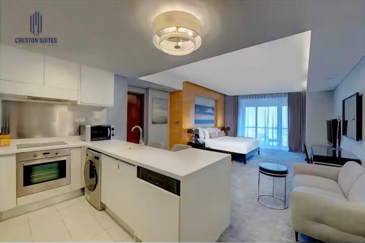 A modern hotel room with a kitchenette, a bed, a sofa, and a large window.