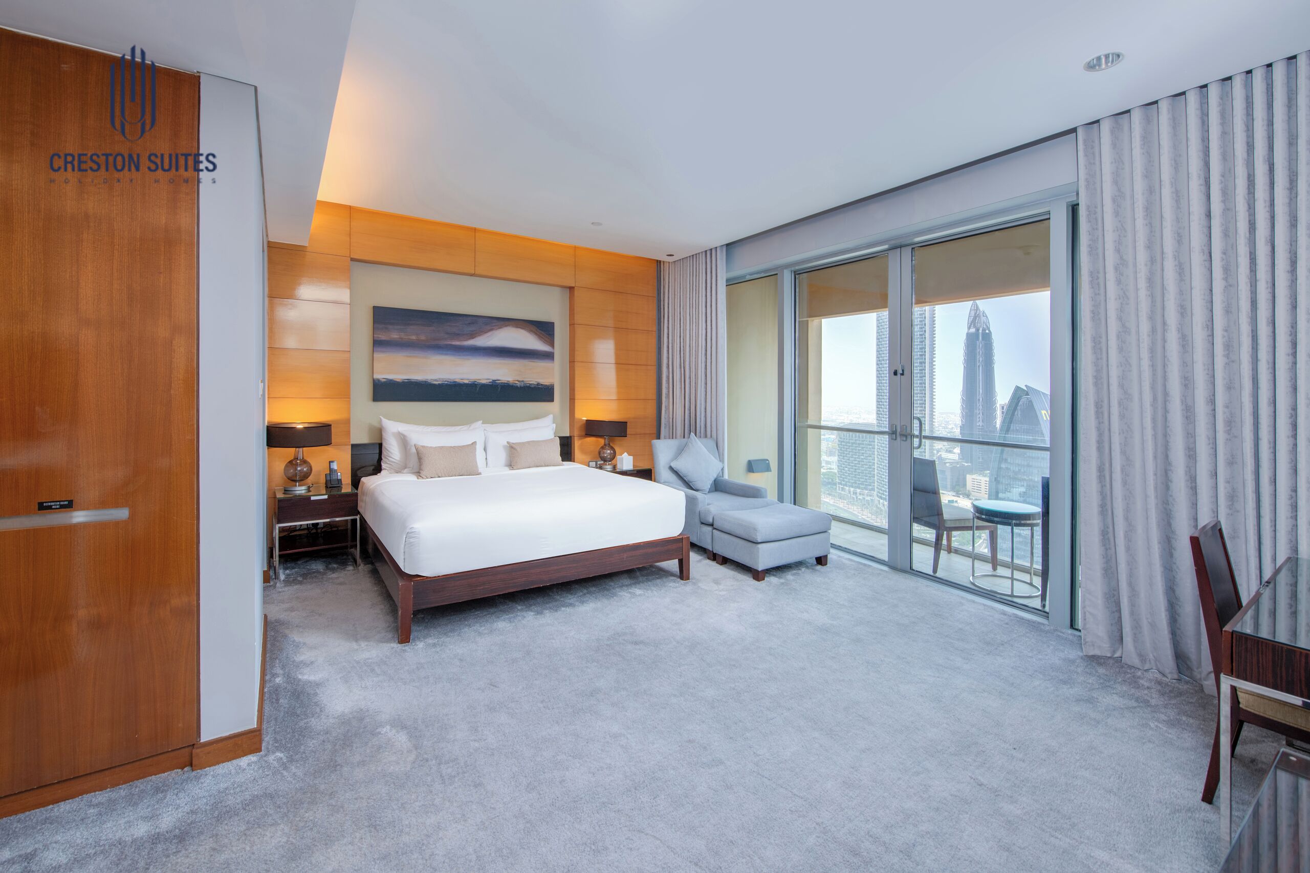 A hotel room with a large bed, a seating area, a view of the city, and a wooden wardrobe.