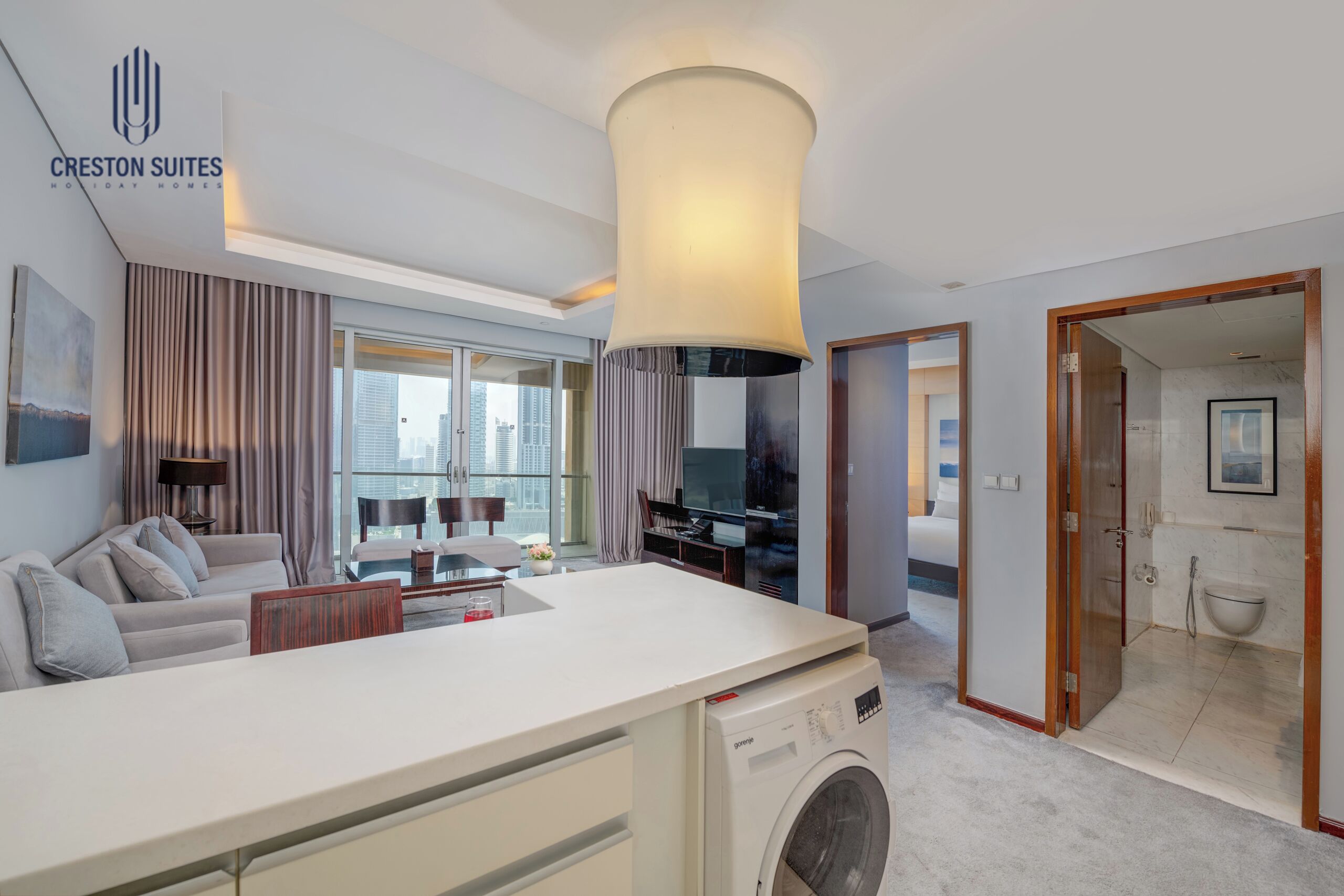 Una sala moderna con sofá, mesa de centro y lavadora. Se ve una ventana grande con cortinas, un televisor y un comedor.