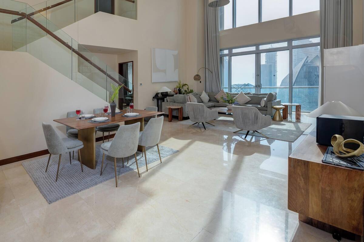 A spacious living area with a dining table, seating, a staircase, and large windows offering a city view.