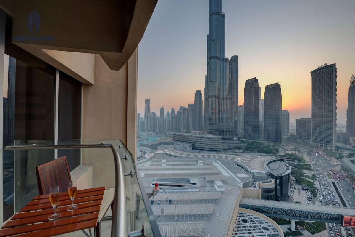 A balcony with a table and two glasses of wine, overlooking a cityscape with high-rise buildings and a large circular structure.