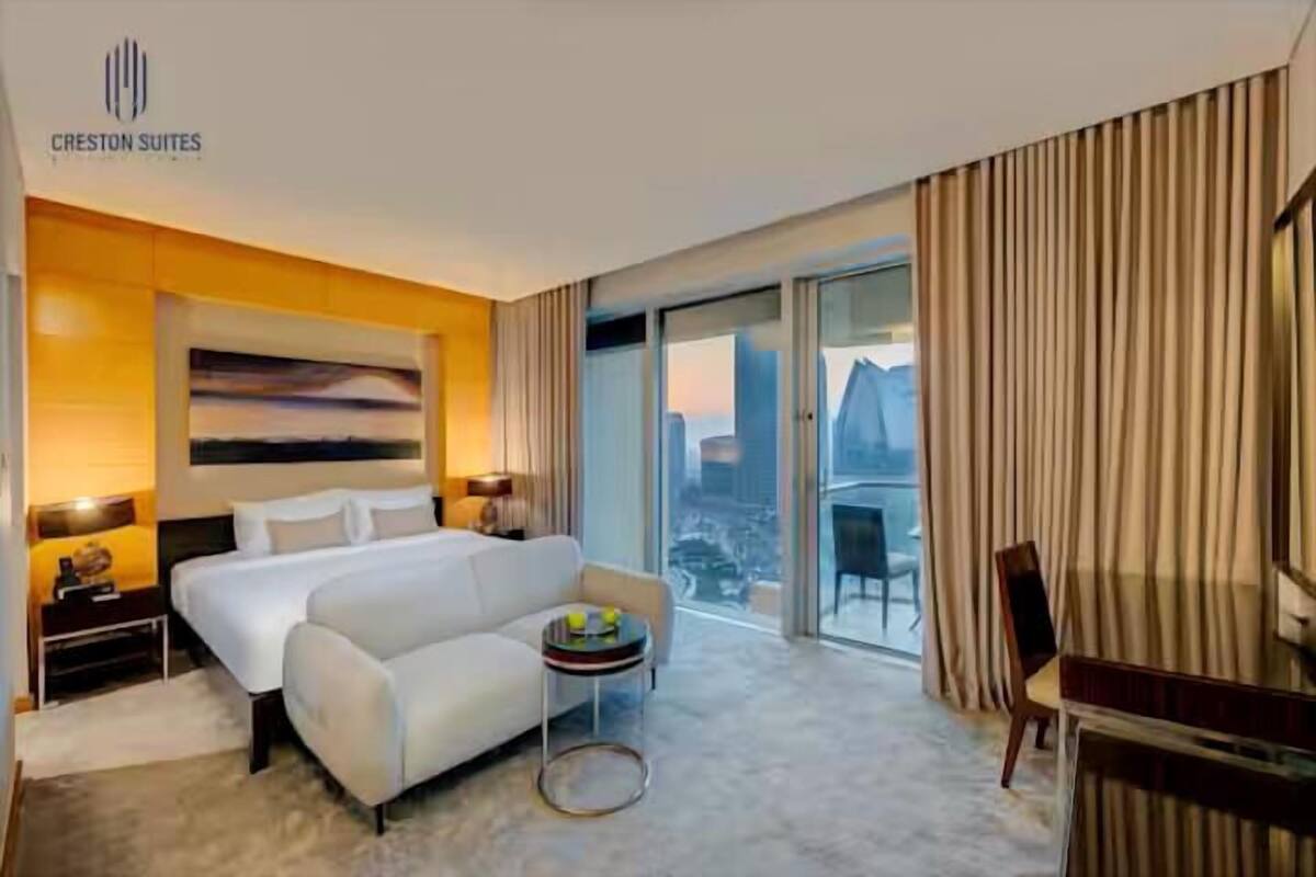 A hotel room with a bed, a small table with a fruit bowl, a desk, and a chair. There is a large window with curtains, and a painting on the wall.
