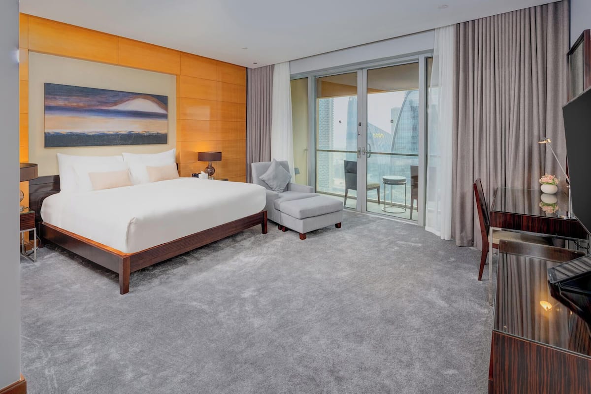 A hotel room with a large bed, a TV, a desk, and a balcony with a city view.