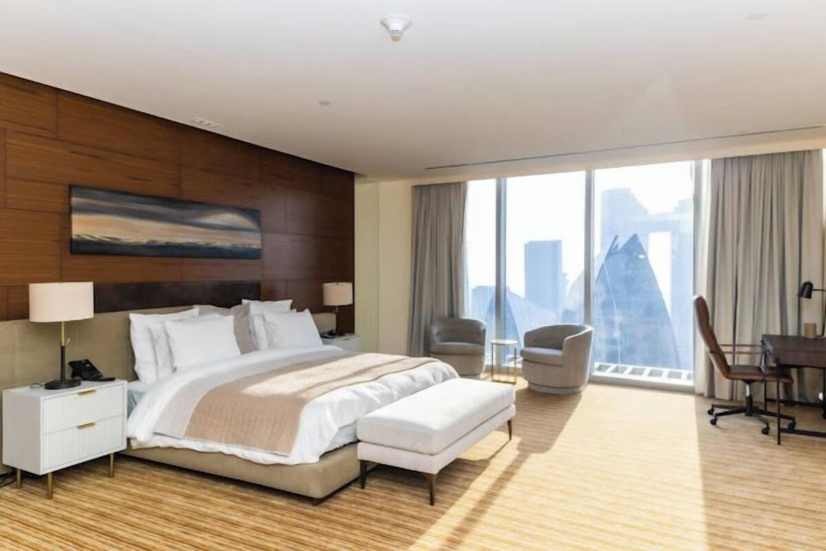 A modern hotel room with a large bed, a desk, and a chair. There is a city view through the window.