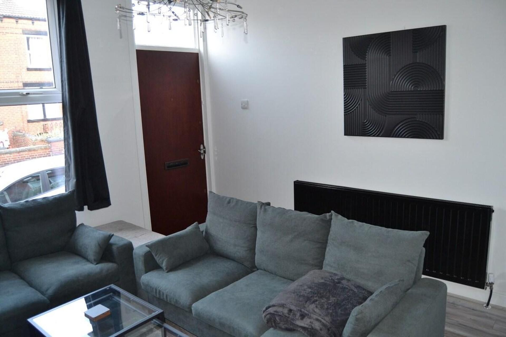 Un salon avec un canapé gris, une table basse en verre et un radiateur. On y trouve une porte et une fenêtre avec des rideaux.