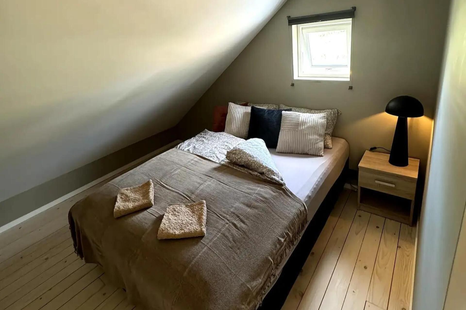 A bedroom with a sloped ceiling, a bed with a beige bedspread and pillows, a nightstand with a lamp, and a window providing natural light.