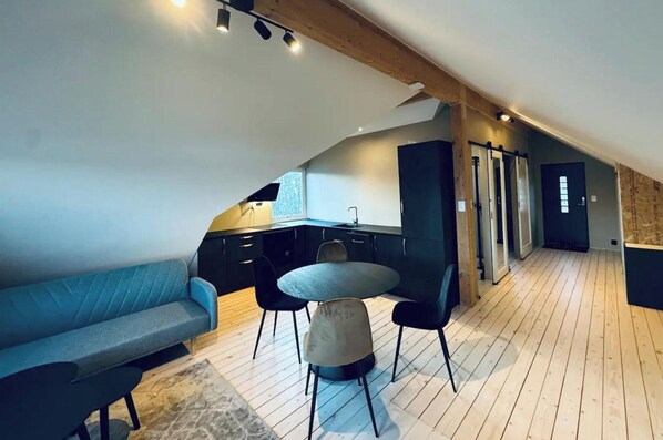 Een loft-achtige woonruimte met een keuken, een eettafel met stoelen, een bank en een trap naar een hoger gelegen niveau.
