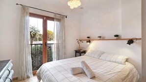 Un dormitorio con una cama grande, un balcón con cortinas, una mesita de noche con lámpara y una repisa con pequeñas plantas.