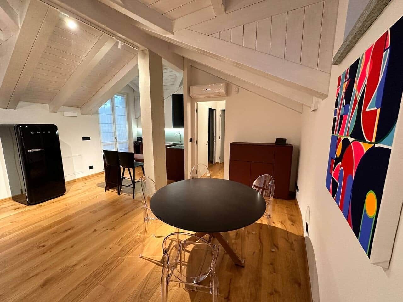 Ein modernes Interieur mit Holzboden, einem runden Tisch mit Stühlen, einer Küchenzeile und einem farbenfrohen, abstrakten Gemälde an der Wand.