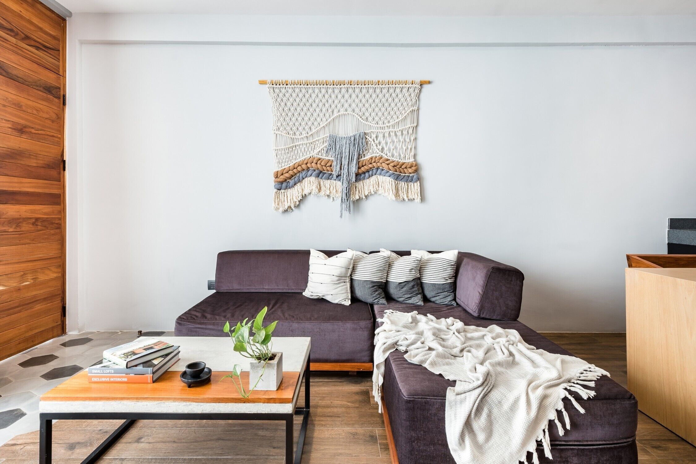 Una sala moderna con un sofá oscuro, una mesa de centro de madera y un colgador de macramé en la pared.