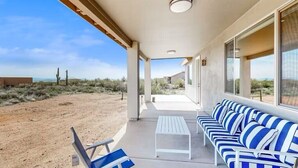 Un patio al aire libre con mesa blanca y sofá azul a rayas, ventanas amplias y vista a un paisaje desértico.