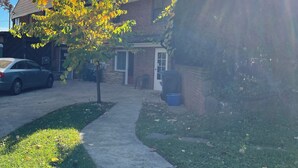 A residential driveway with a parked car, a house with a brick facade, and a tree with yellow leaves. - Room with Two DB Beds, Bathroom, Fridge, MW, TV, Wifi - 1 guest 64, 2 guests 120 (Silver Spring)