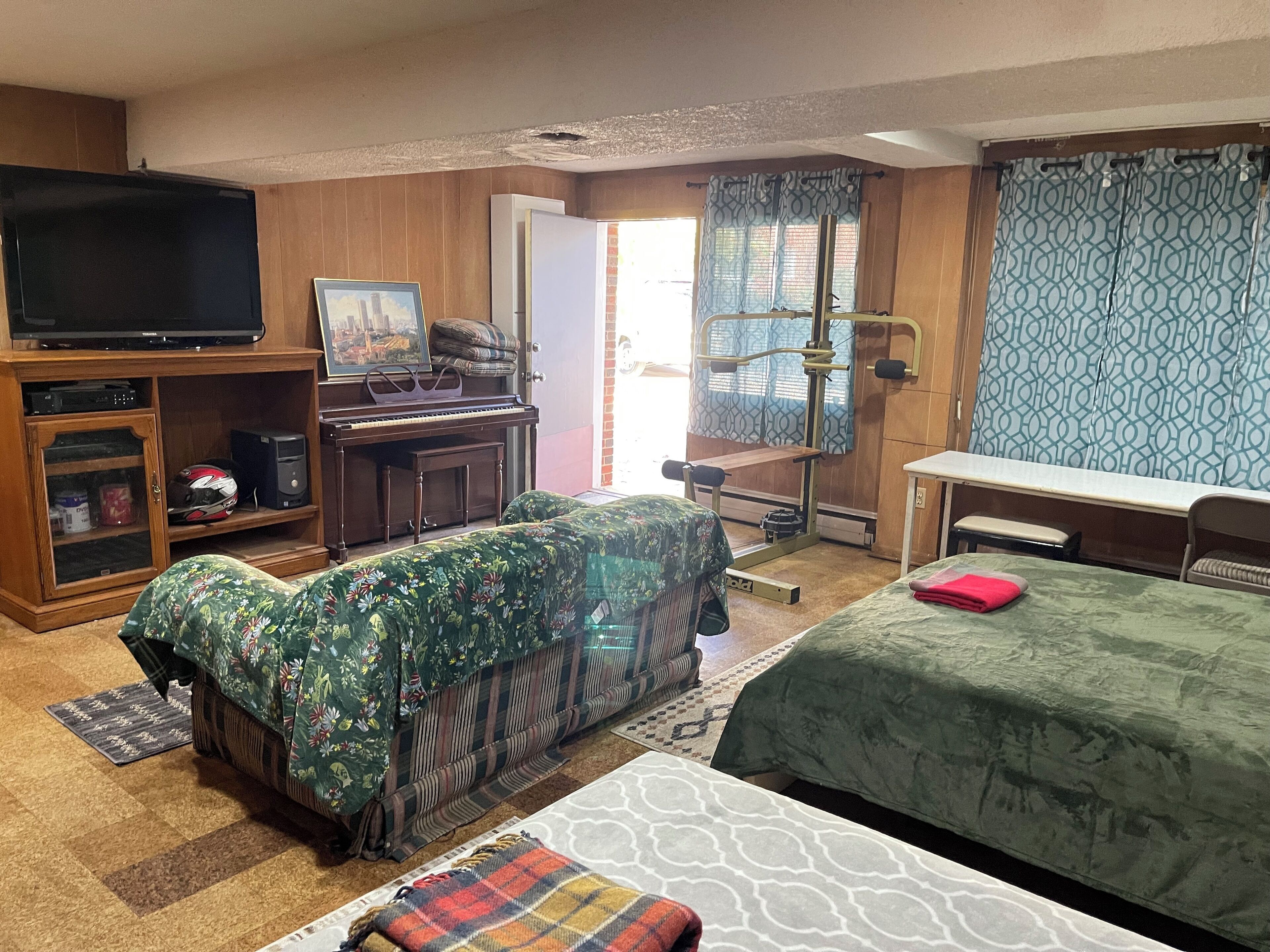 A room with a bed, a desk, a television, a piano, and a window with curtains.