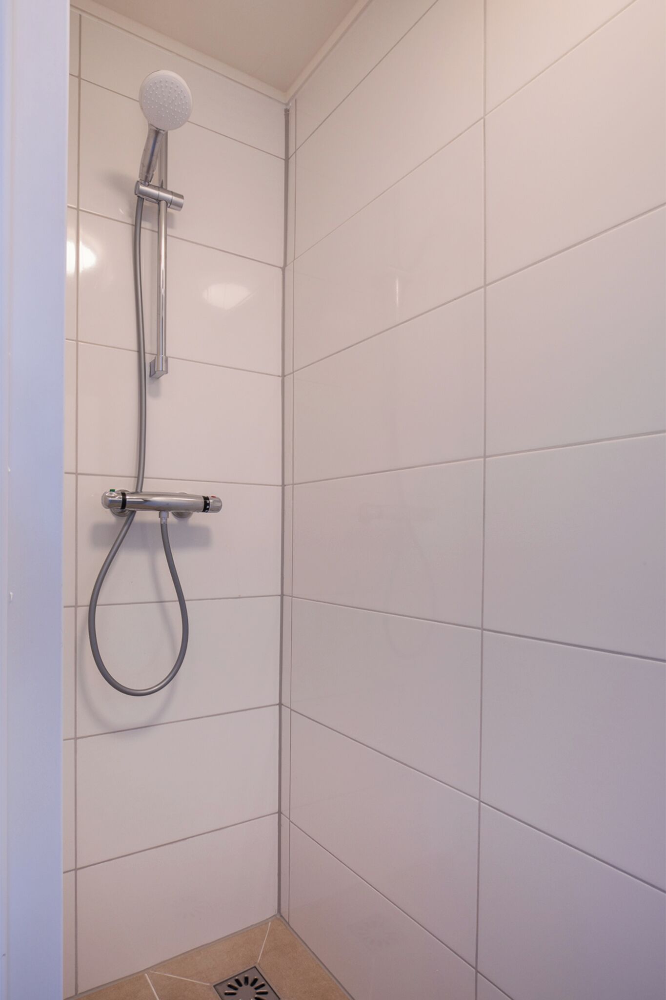 A shower with a handheld showerhead and a wall-mounted control valve in a corner with white tiled walls.