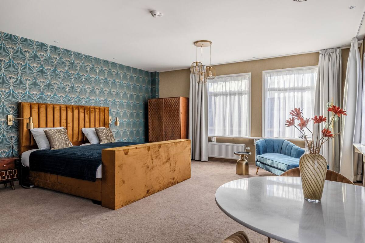 Une chambre à coucher avec un lit, une tête de lit en bois, un canapé bleu, une table ronde et un vase rempli de fleurs.