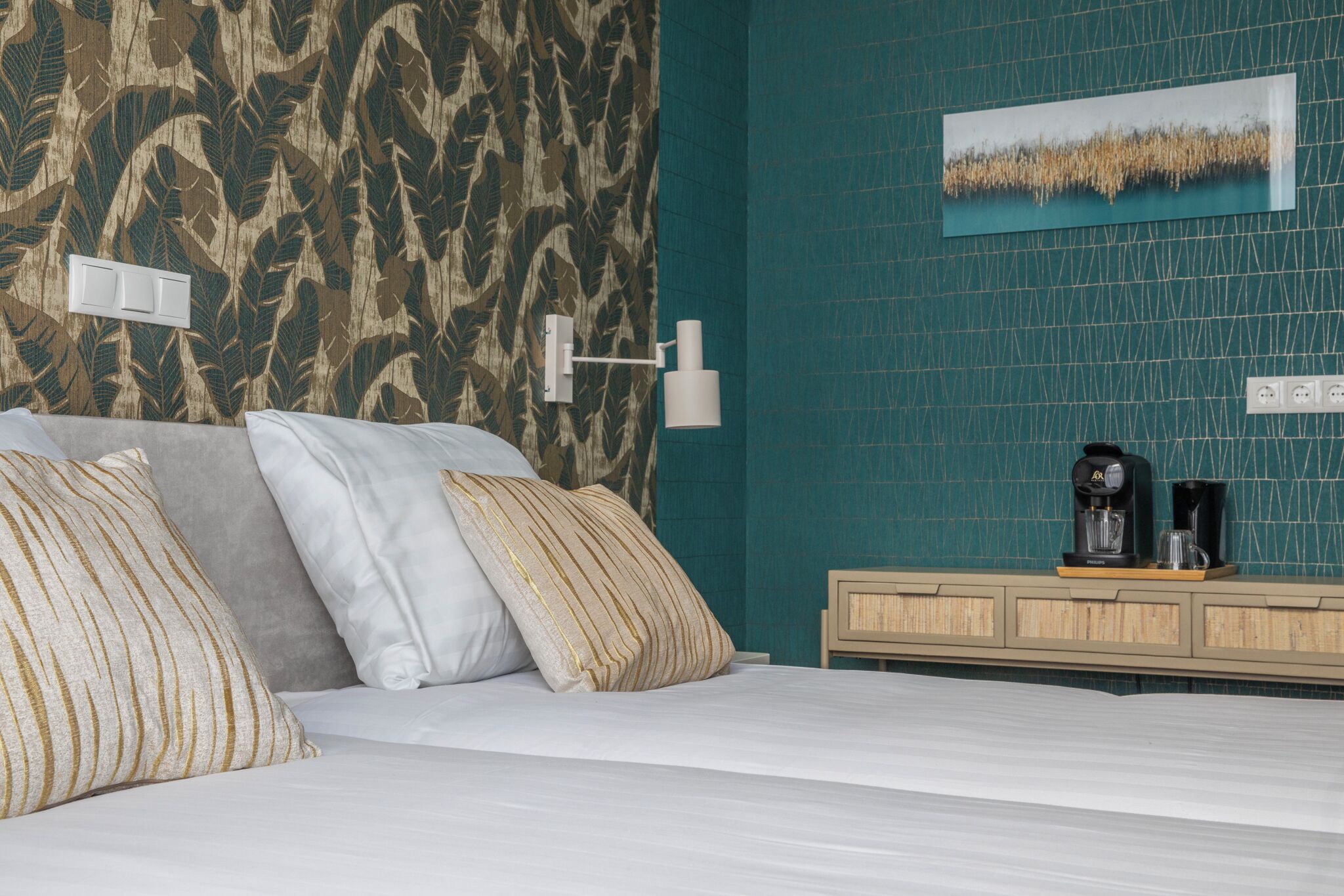 A bedroom with a bed, pillows, a nightstand, a coffee maker, and a wall with a patterned wallpaper and a painting.