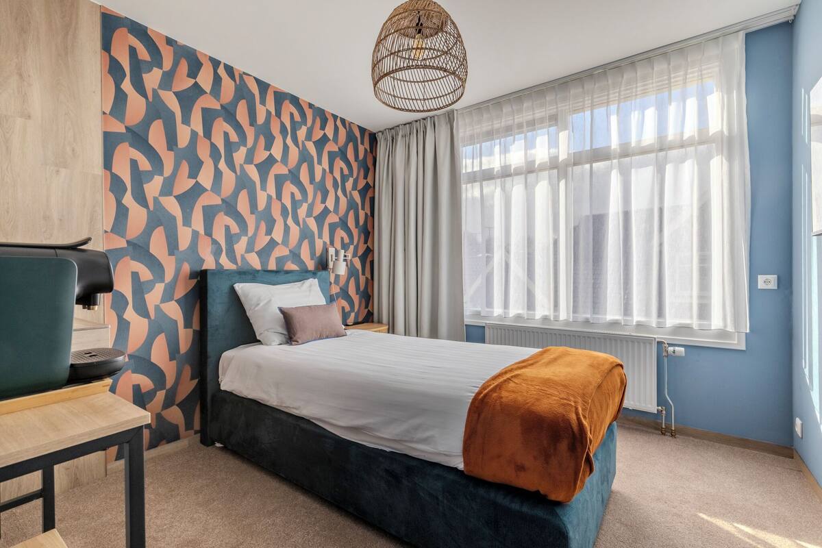 Une chambre d’hôtel moderne avec un grand lit, un bureau, une chaise et une fenêtre avec des rideaux légers.