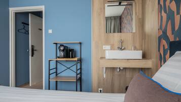 A bedroom with a bed, a wooden dresser, a small table with a kettle, and a bathroom with a sink and mirror.